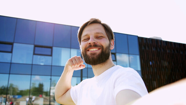 Cheerful young man with clenched fist taking selfie and smiling cheerfully outdoors, first person view. Happy bearded guy celebrating some successful news.