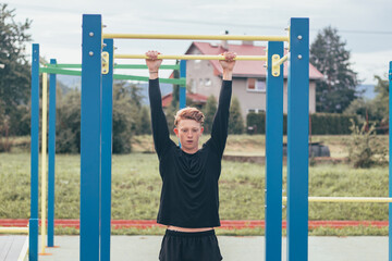 Goal-oriented blond boy builds the strength of his own body on the workout field during the summer. Training triceps, biceps, shoulders and abdomens. Crosfit exercise pull-ups