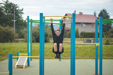 Goal-oriented blond boy builds the strength of his own body on the workout field during the summer. Training triceps, biceps, shoulders and abdomens. Crosfit exercise pull-ups