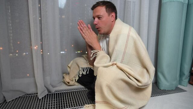 happy man wrapped in a blanket sits on a heating convector and warms his hands on the grates of a warm heater