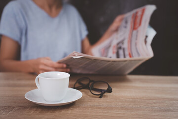 A cup of coffee,glasses on the wood table and woman read a newspaper.