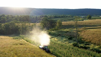 Aerial view of tractor spraying corn in a field with pesticides at sunrise in the mountains.