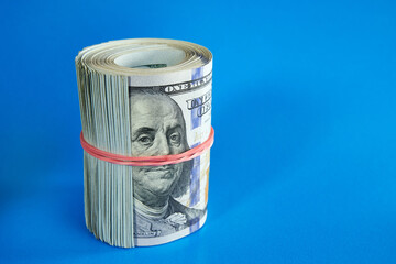 Many hundred dollar bills rolled up with a red rubber band on a blue background.