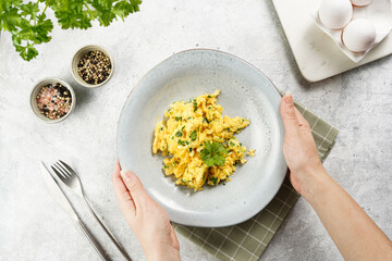 Female hands holding breakfast scrambled eggs with green herbs, parsley in deep grey bowl on grey neutral table, vintage fork and knife, top view - obrazy, fototapety, plakaty