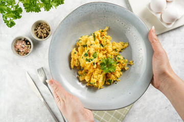Female hands holding breakfast scrambled eggs with green herbs, parsley in deep grey bowl on grey neutral table, vintage fork and knife, top view