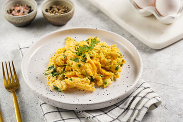 Breakfast scrambled eggs with green herbs, parsley in white plate on grey neutral table, golden fork and knife