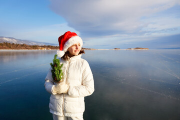 Fototapeta premium A girl in a Santa Claus hat holds a small Christmas tree in her hands. A girl stands on the ice of the frozen Lake Baikal. Celebrating New Year and Christmas.