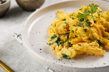 Breakfast scrambled eggs with green herbs, parsley in white plate on grey neutral table, golden fork and knife