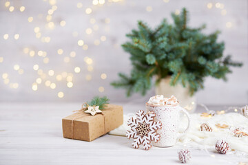 Christmas greeting card. A mug of cocoa or hot chocolate with marshmallows, a gift in kraft paper, branches of a fir tree on a beautiful bokeh background.