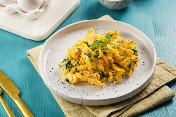 Breakfast scrambled eggs with green herbs, parsley in white plate on vivid blue wooden table, golden fork and knife
