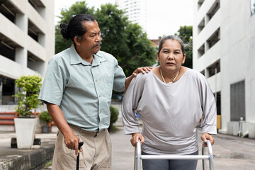 Senior wife with knee or leg injury, using walking aid with supporting old husband, concept of supporting senior couple or suffering aging society with difficulties