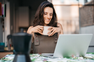 A young Mediterranean brunette woman smiles while checking her laptop and sipping a cup of coffee or tea while working from the terrace of her house.