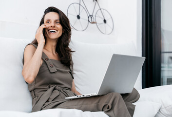 A young Mediterranean brunette woman smiles brightly as she makes a phone call. She works with her laptop on the white sofa on her terrace.