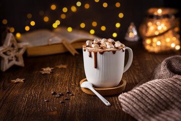 Hot chocolate with marshmallows in a white cup on a wooden table with a book and a garland in the background.