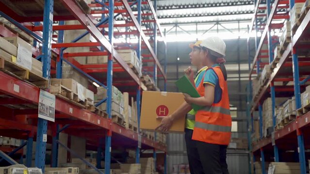 4K, Slow motion, Two Asian male and female employees, counting the boxes inside the warehouse, sorting the goods on the shelf, they both wear helmets and reflective green vests for their own safety.
