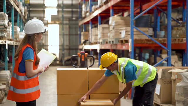 4K, Female Chief Clerk at the warehouse waiting for a colleague Load crates of goods and arrange on carts. Prepare to deliver to customers order in. Wear helmet and green reflective vest for safety.