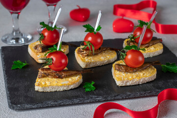 Canape in the form of hearts with sprats, cheese, egg and cherry tomatoes on slate plate on light gray background, idea for Valentine's Day