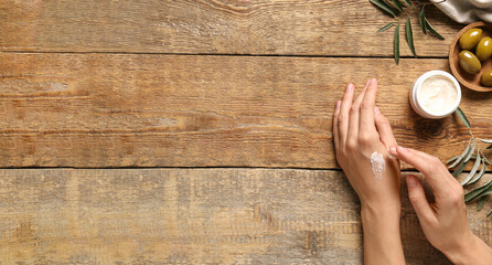 Woman applying cream with olive oil extract onto her hands on wooden background with space for text, top view