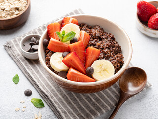 Chocolate oatmeal porridge served with chocolate chips, fresh strawberry and banana slices. Close up view. Homemade breakfast. Healthy eating concept. Vegetarian food. Light grey background.