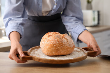 Woman putting board with fresh bread onto table in kitchen, closeup
