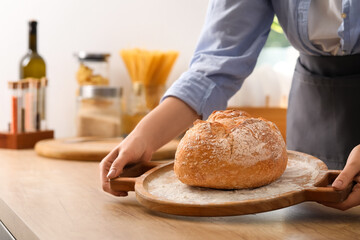 Woman putting board with fresh bread onto table in kitchen, closeup