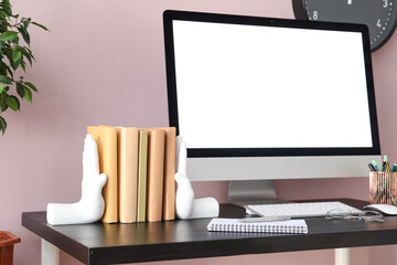 Stylish holder with books, computer and eyeglasses on table near pink wall
