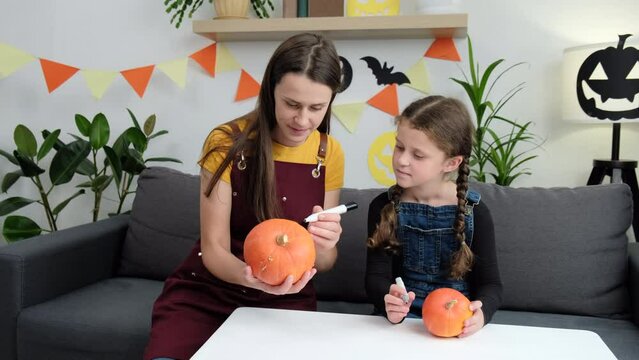 Cheerful young mother and cute daughter child sitting on comfy couch painting small orange pumpkins spooky pumpkin and joking, family preparing for celebration of Halloween. Autumn holiday concept