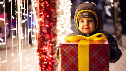 Happy child in a red Santa Claus hat with wrapped Christmas gift near shining light bulbs in winter evening. The concept of Christmas and New Year