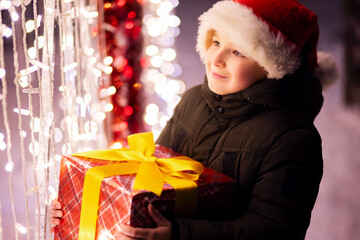 Fototapeta na wymiar Happy child in a red Santa Claus hat with wrapped Christmas gift near shining light bulbs in winter evening. The concept of Christmas and New Year
