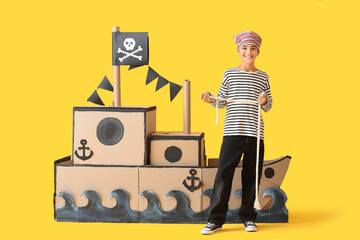 Little boy dressed as pirate with rope and cardboard ship on yellow background