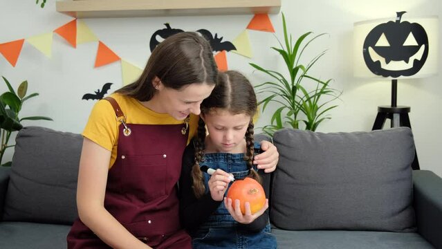 Happy loving young mother teaches cute daughter painting Halloween pumpkin sit together on cozy couch at home, smiling parent and little kid sitting at table and preparing handmade decorations