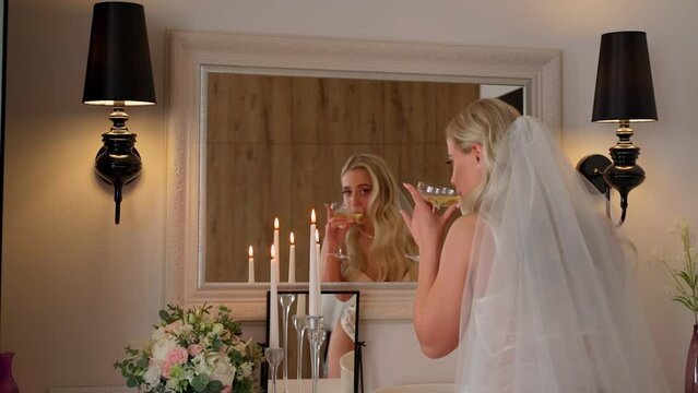 Attractive bride in lingerie looking in a mirror dring champagne in living room. Beautiful caucasian young adult girl prepare to wedding day. Sensuality and femininity of women on marriage event