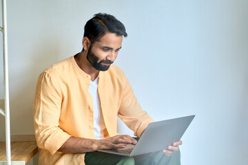 Smiling indian man professional user sitting on stairs typing on laptop device, using computer for online work or remote learning, elearning, surfing internet digital technology, relaxing at home.