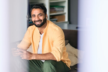 Happy ethnic indian man sitting at home on couch wearing earbuds using mobile phone looking at camera enjoying listening music, podcast or having fun relaxing chatting using smartphone and earbuds.