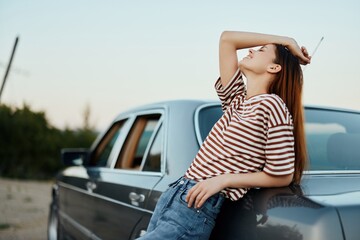 A young woman in a striped T-shirt and jeans stands by her car resting from driving, leaning on her car beside the road and smiling 