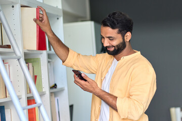 Happy smiling indian ethnic man using phone standing at home taking book from library shelves. Male reader checking bookstore app on smartphone searching ebooks in virtual mobile bookshop.