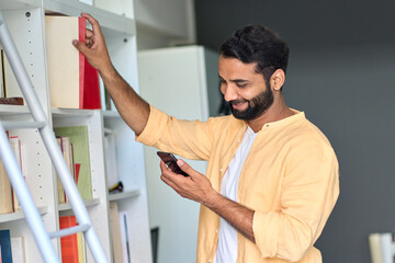 Happy smiling indian ethnic man using phone standing at home taking book from library shelves. Male...
