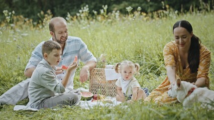 Family with dog having picnic in field. Happy parents and kids eating watermelon and petting obedient dog while having picnic in grassy meadow on summer weekend day in nature
