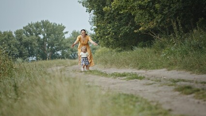 Mom and daughter running on countryside road. Mother in yellow dress chasing happy daughter running on countryside road then stopping and telling secret on weekend day in nature