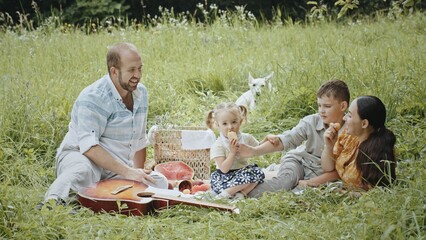 Happy relaxed young family posing in sunlight on summer picnic outdoors. Smiling Caucasian man, woman, little boy and infant looking at camera sitting on blanket on sunny meadow.