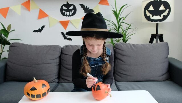 Cute little girl preparing for Halloween, painting drawing scary face on pumpkin while sitting at white table in living room at home, smiling child making jack-o-lantern. Holiday decoration concept