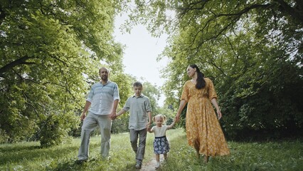 Family strolling near trees in countryside. Ground level zoom out view of happy parents and kids holding hands and swinging little girl while walking on path amidst lush trees on sunny weekend day in