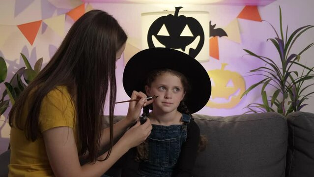 Close up of cute young mother painting face little daughter child for Halloween celebration, family sitting together on cozy couch in dark living room decorated spooky paper black pumpkins and bats