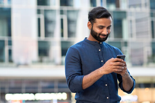 Smiling indian business man, eastern businessman holding cell phone using smartphone mobile apps texting message, surfing social media tech standing in urban city on modern street outdoors.