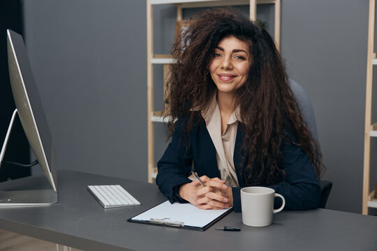 Cute happy friendly tanned adorable curly Latin businesswoman in jacket enjoy at work project smile at camera in office. Copy space Mockup Banner. Corporation leader lady uses modern computer in work