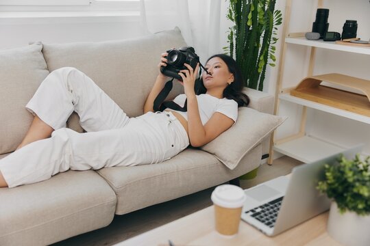 Asian woman photographer lying at home on couch and looking at photos in camera, freelance photographer business for self