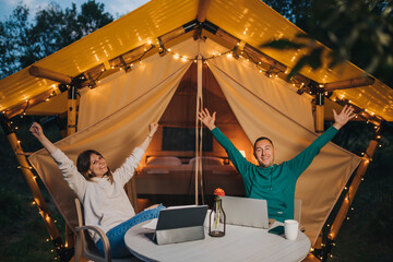 Happy family couple freelancers working laptop on a cozy glamping tent in summer evening. Luxury camping tent for outdoor holiday and vacation. Lifestyle concept