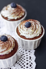 three chocolate muffins with delicate butter cream and blueberries stand on a lace napkin on a dark table. delicious dessert.
