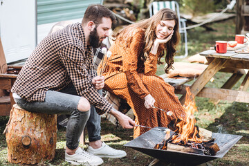 Young beautiful loving couple travelling across country in the van. Millennial man and woman in a travel camper. Cozy atmosphere, vacations vibe. Drinking tea, cooking marshmallow on a campfire
