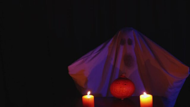 celebrating halloween home party. ghost costume from white sheet and spooky creepy face moving vowing hands wings above pumpkin and two burning flame candles in night dark background. concept scary